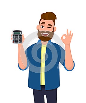 Young man showing or holding digital calculator device in hand and gesturing, making okay or OK sign while winking eye. Good.