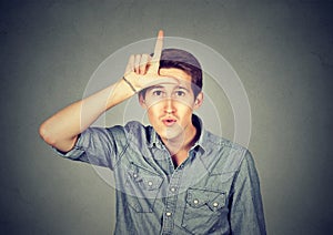 Young man showing loser sign on forehead, looking with disgust