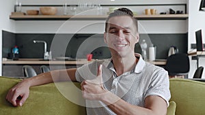 Young man showing thumb up sign while sitting on sofa at home. Smiling male shows gesture of approval while sitting on