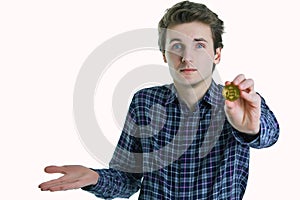 Young man shrugs his shoulders, not understanding what to do with bitcoin in his hand isolated on white background.