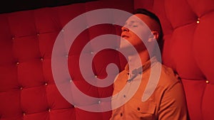 young man singing on a sofa in red room of nightclub. Shooting music video