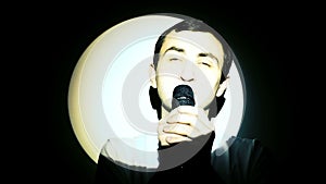 A young man sings into a microphone in the white light beam