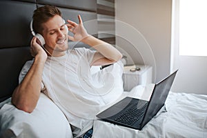 Young man sit in bed early morning. He enjoying listen to music through headphones. Positive guy has laptop on bed