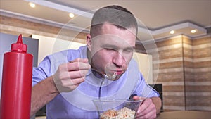 Young man sit in kitchen and eat pasta with ketchup or sauce. Guy takes some food with fork and put it in mouth. Eating