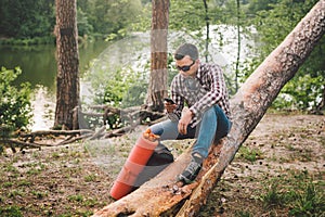 Young man sit on truck in forest and using mobile phone. man traveler sits on large fallen tree, holding phone in hand. theme