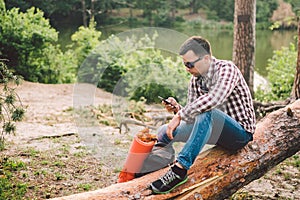 Young man sit on truck in forest and using mobile phone. man traveler sits on large fallen tree, holding phone in hand. theme