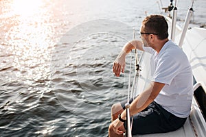 Young man sits at adge of yacht board and looks forward on sun. He leans on railing. Guy is calm and peceful. He is photo