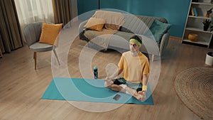 A young man sits cross legged in a lotus position with his eyes closed on a sports mat next to a smartphone. A man in a