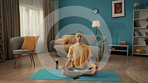 A young man sits cross legged in a lotus position with his eyes closed on a sports mat next to a smartphone. A man in a