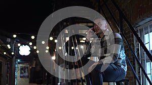 A young man sits on a fire escape and talks on the phone. A man on the stairs of a narrow street late at night.