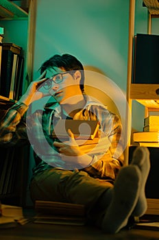 An young man sits on the floor between bookshelves in the evening, wearing glasses and holding a stack of books with a shocked