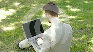 Young man sits on a grass in the park and taping on laptop computer