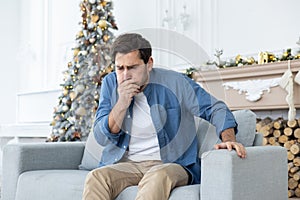 A young man sits at home on the couch during the New Year holidays and feels very nauseous, covers his mouth with his