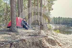 Young man sits near a tent in the forest on a high bank of the river