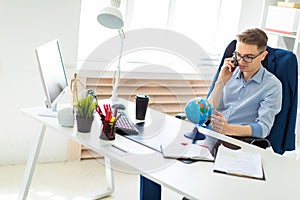 A young man sits in the office at a computer desk, talking on the phone and looking at the globe.