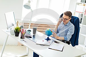 A young man sits in the office at a computer desk, talking on the phone and looking at the globe.