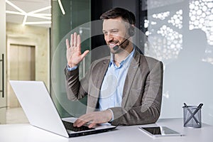 A young man sits in the office at a desk in a headset and works on a laptop, talking and greeting to the camera online