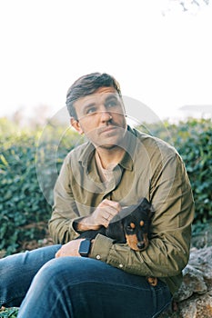 Young man sits in a park and strokes a black puppy on his knees while looking away