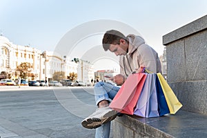 Young man sits with phone in his hands next to multi-colored shopping bags