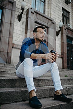 Young man sits and ponders on stairs with cup of coffee near building