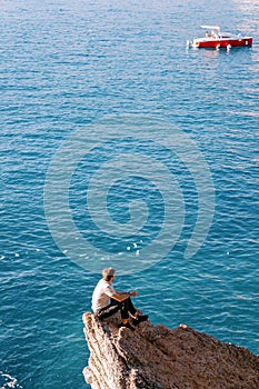 Young man sits on a rock ledge and looks at a boat sailing on the sea. Back view