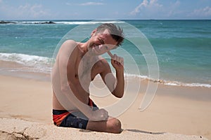 A young man sits on a sandy beach and tries to clear his ear from salt water.