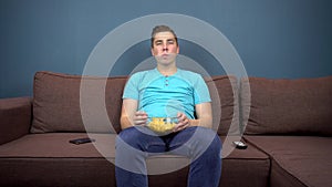 A young man sits on a sofa eating chips and watching TV. The view from the TV. Looking at the camera