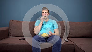 A young man sits on a sofa eating chips and watching TV. The view from the TV. Looking at the camera