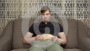 A young man sits on a sofa and touches his fat belly. Gained excess weight in quarantine.