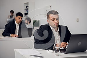 Young man sits at a table and works with laptop, colleages sit on the background