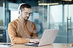 A young man sits at a workplace in the office, works, studies on a laptop with headphones. Listens to the webinar, music