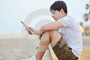 Young man sitting on beach reading ebook