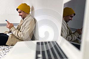 Young man sitting in bed and using mobile while laughing