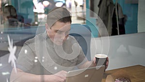 Young man sitting behind the glass in a cafe with a laptop. The man drinks coffee and works.