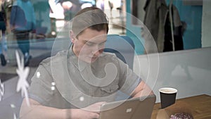 Young man sitting behind the glass in a cafe with a laptop. The man drinks coffee and works.