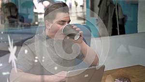 Young man sitting behind the glass in a cafe with a laptop. The man drinks coffee and works.