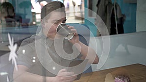 A young man is sitting behind a glass in a cafe with a tablet in his hands. The man drinks coffee and works.