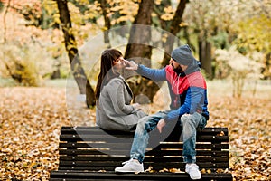 A young man is sitting on a bench in an autumn park with his beloved girlfriend and gently touches her nose with his