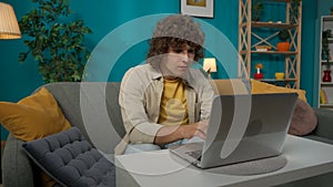 Young man sitting on the couch in the living room and typing something on his laptop with serious face