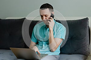 Young man sitting on the couch, speaking on the phone and working on laptop