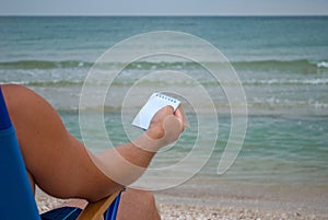 Young man sitting on a deckchair on the shore, holding a notebook for notes in the cage, resting sunbathing