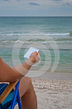 Young man sitting on a deckchair on the shore, holding a notebook for notes in the cage, resting sunbathing looks at the sea