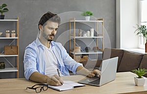 Young man sitting at desk with laptop computer, having online class and taking notes