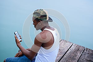 Young man sitting at the edge of a pier and looking at his mobile phone attentively