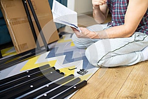 Young man sitting on the floor and reading instructions for assembling furniture at home.