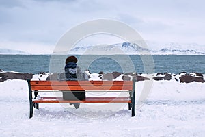 young man sitting in front of a lake on a bench at the snow-capped mountains in the background