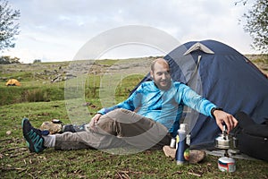 Young man sitting in front of a tent, smiling, heating water on his stove