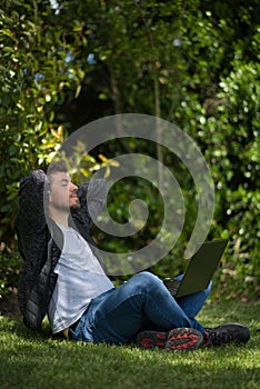 Young man sitting on the grass with his computer on his legs while stretching and relaxing on a park. Natural Environment.