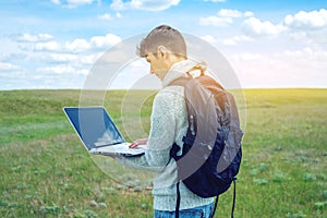 Young man sitting on a green meadow with laptop wireless on the background of blue cloudy sky