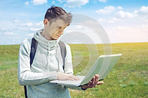 Young man sitting on a green meadow with laptop wireless on the background of blue cloudy sky